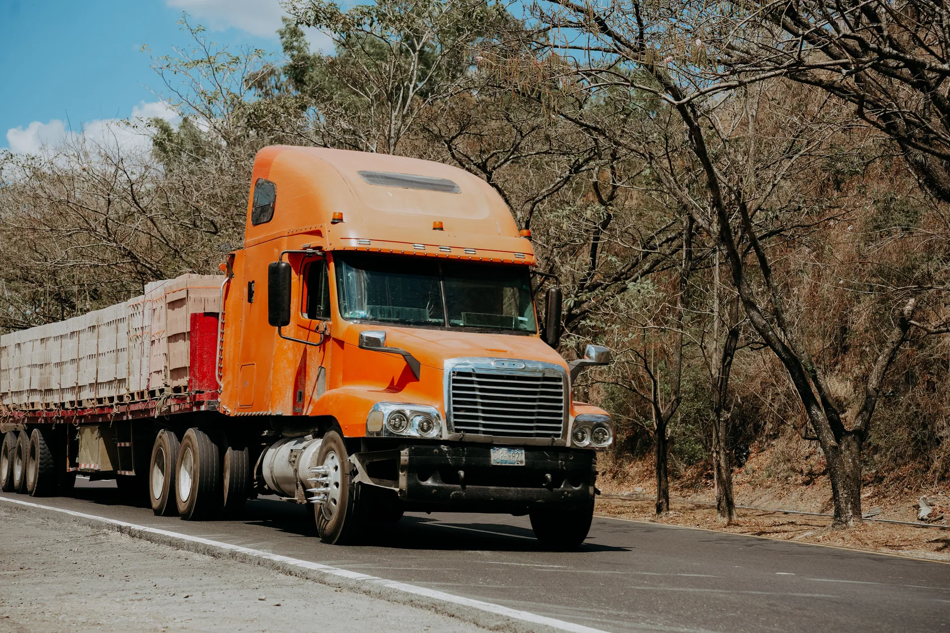 A vibrant orange Freightliner truck drives on a highway through a dry, scenic landscape in El Salvador.