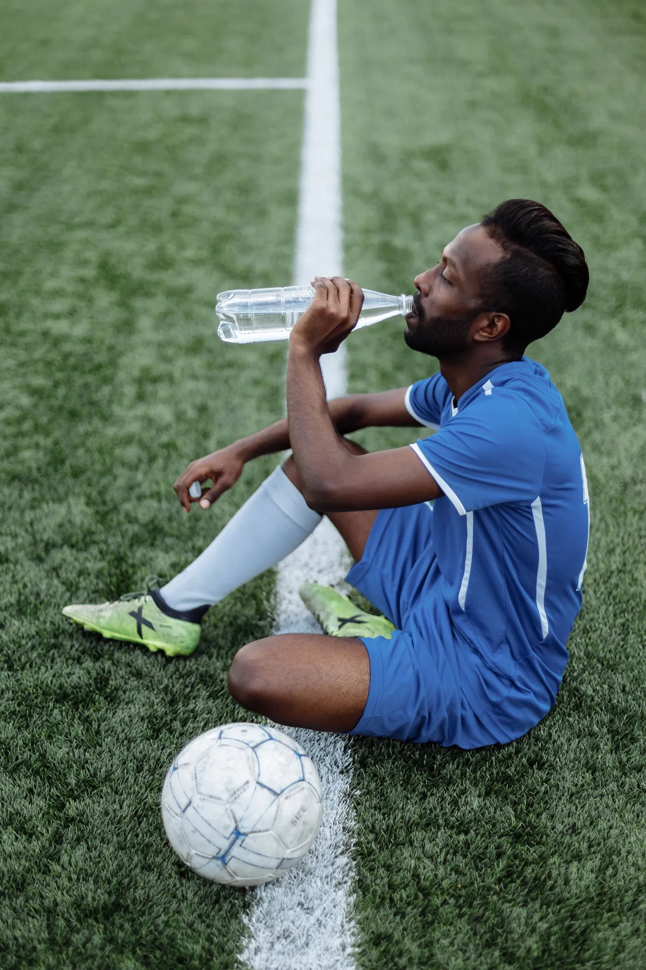 Athlete in blue soccer jersey sitting on grass, hydrating during a break.