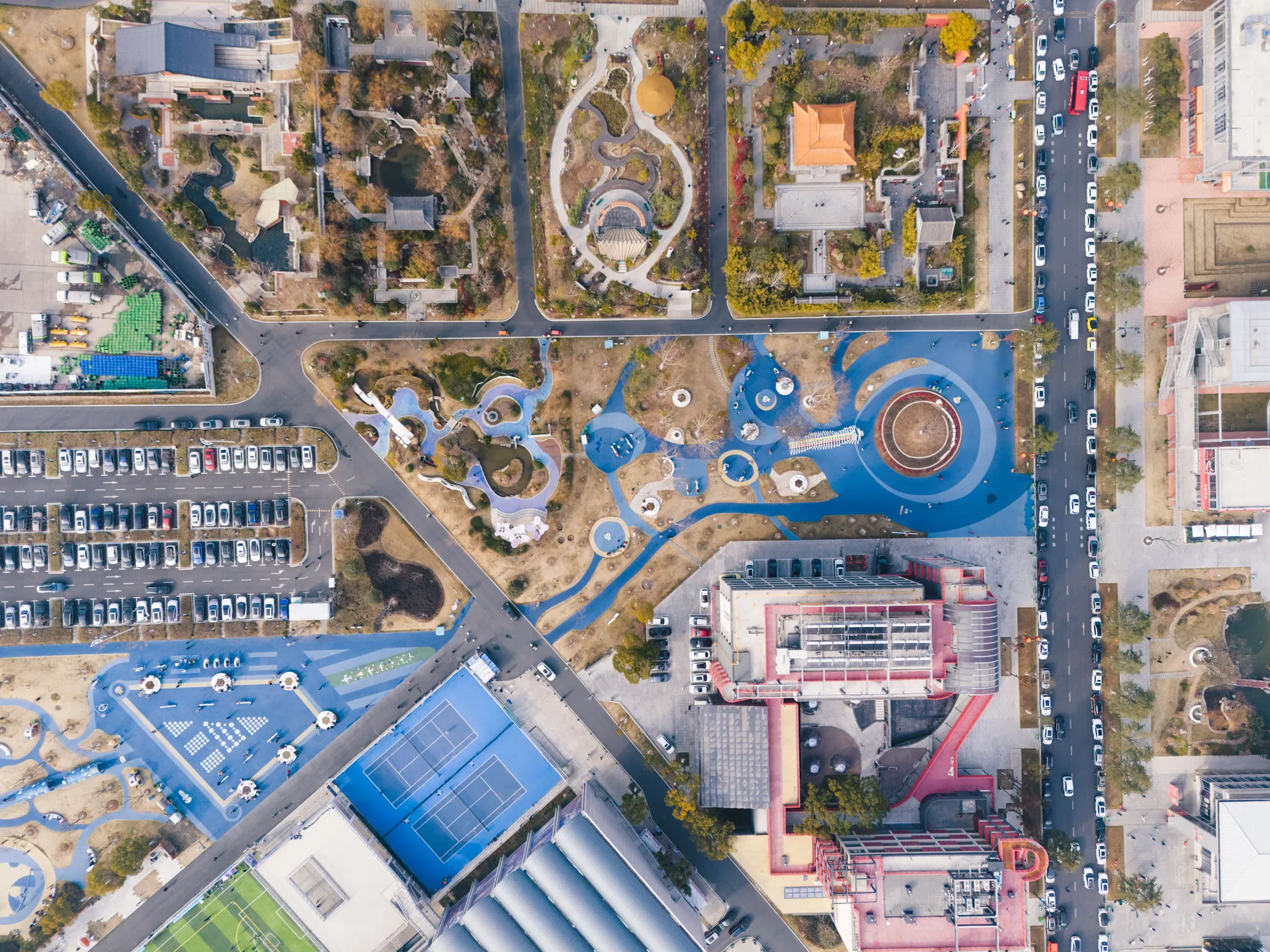 Aerial view of an urban park with playgrounds, parking, and nearby buildings.