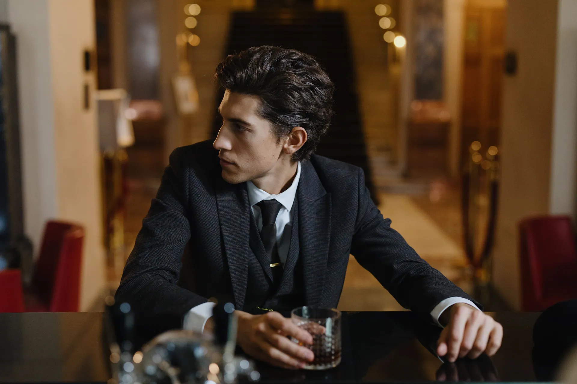 Stylish man in a dark suit sits at a luxurious indoor bar, holding a glass of whisky.