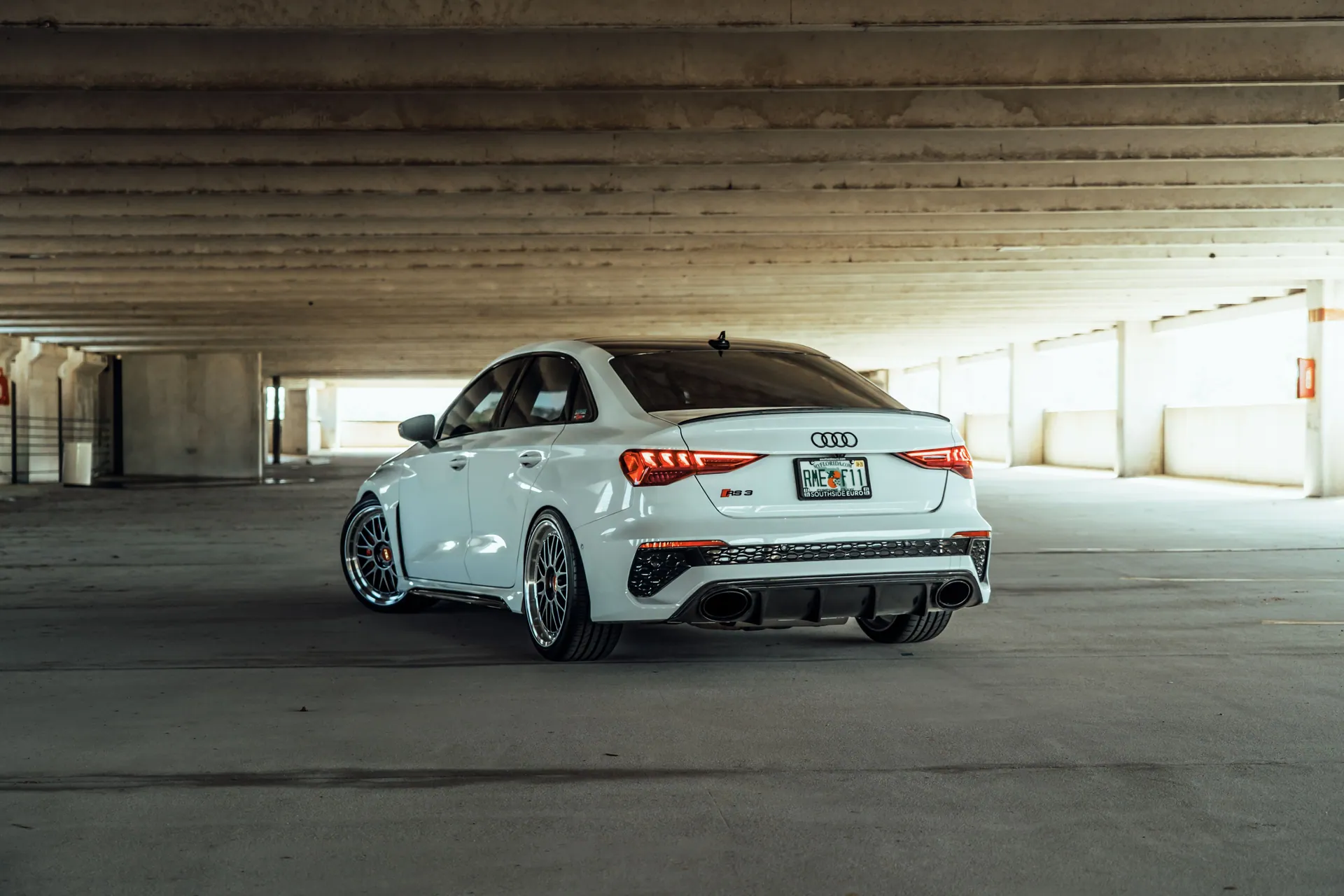 White Audi RS3 parked in a dimly lit, modern, urban parking garage. Rear view accentuates sporty design.