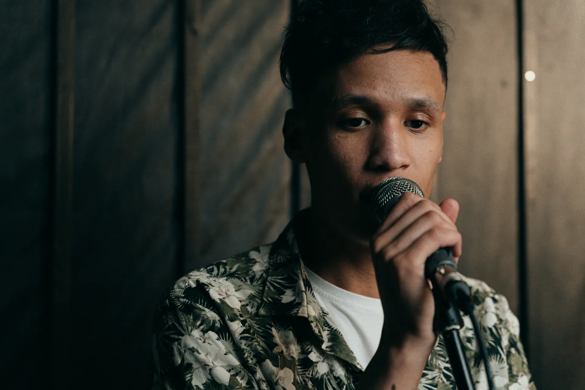 A young man wearing a floral shirt sings into a microphone indoors. The atmosphere is moody and intimate.