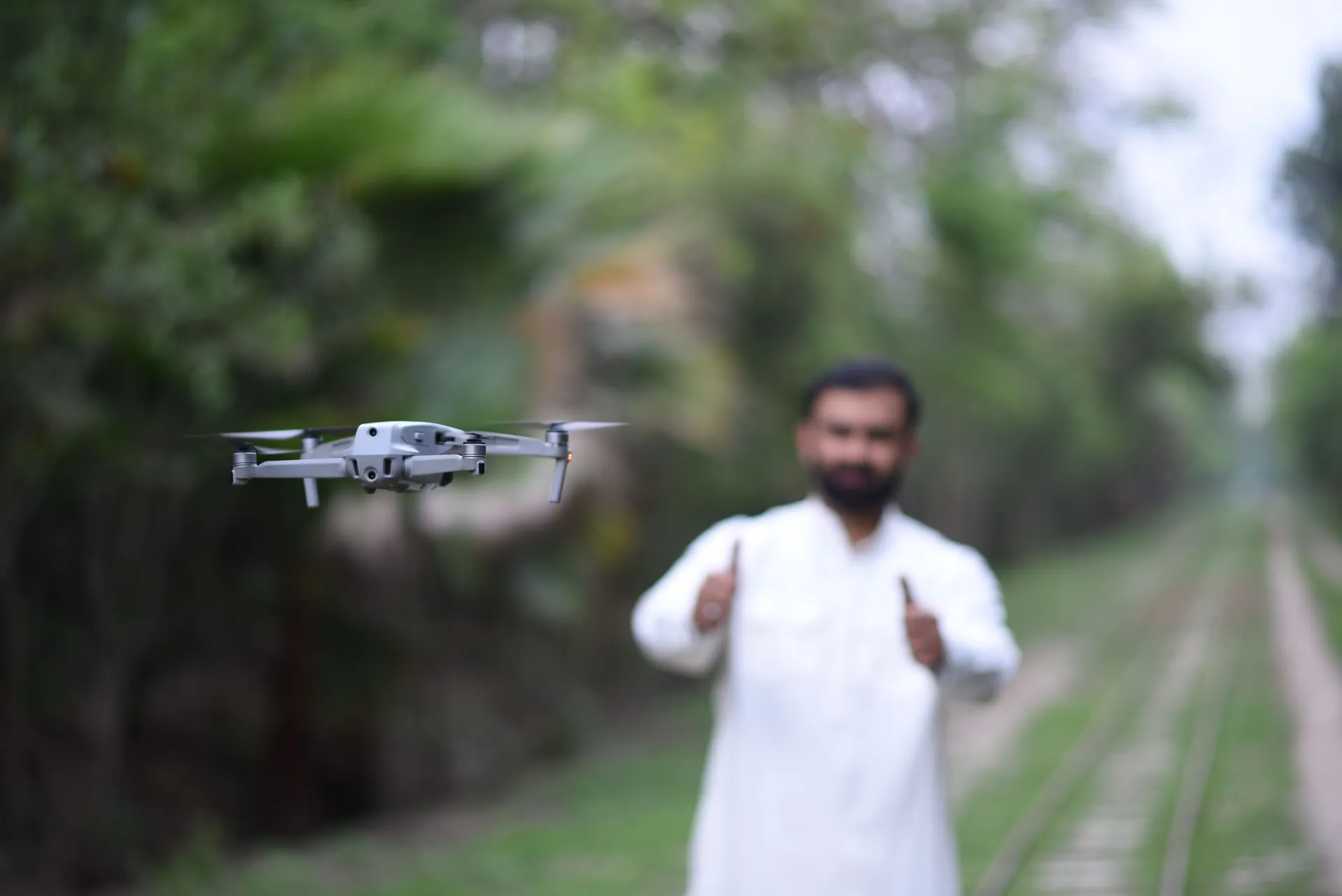 A man gives a thumbs-up while operating a drone in a forested pathway.