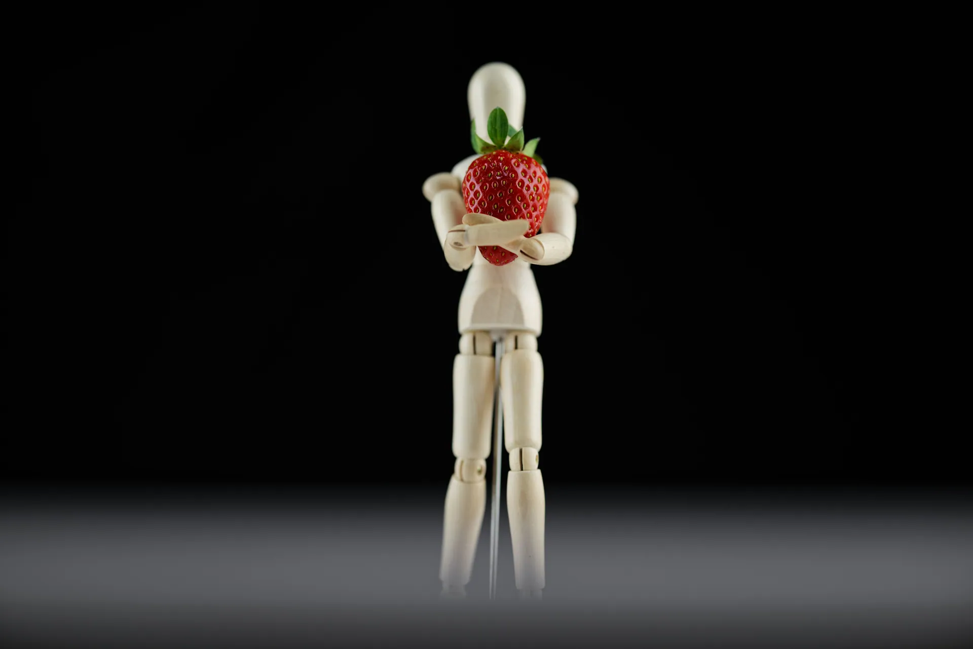A wooden mannequin holding a vibrant strawberry against a black background, captured in a studio setting.