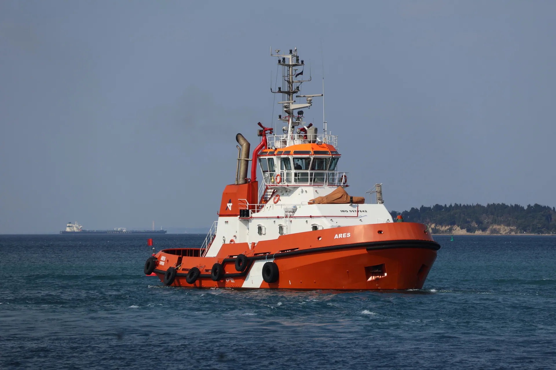 Orange tugboat ARES sailing on calm waters near the coastline on a sunny day.