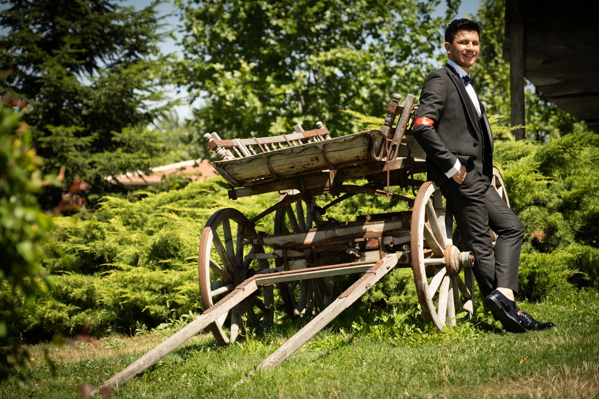 A stylish groom in a black suit poses with a vintage wooden cart outdoors, showcasing rural elegance.