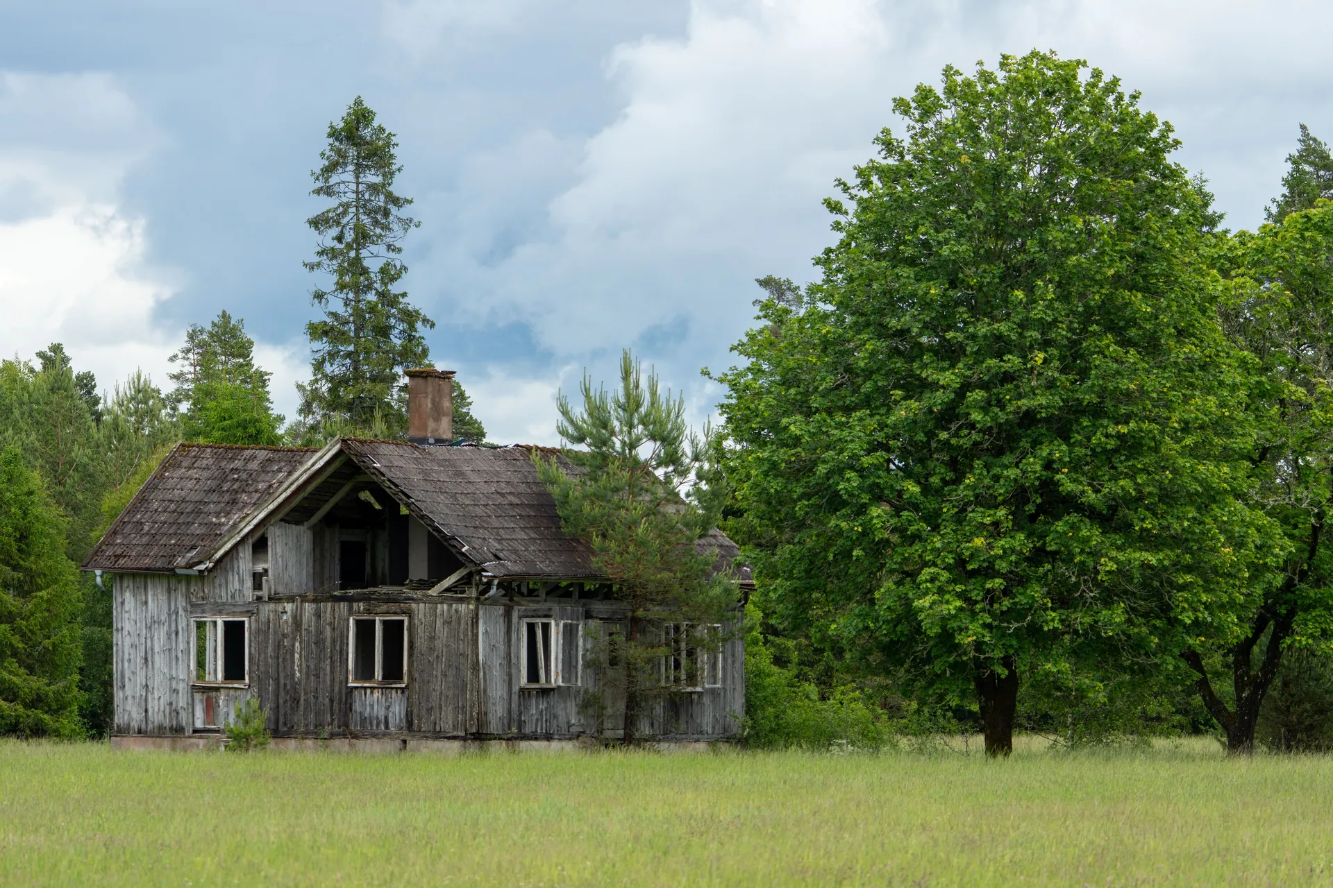 Scenic view of an old, abandoned wooden house in a green field surrounded by trees under a cloudy sky.