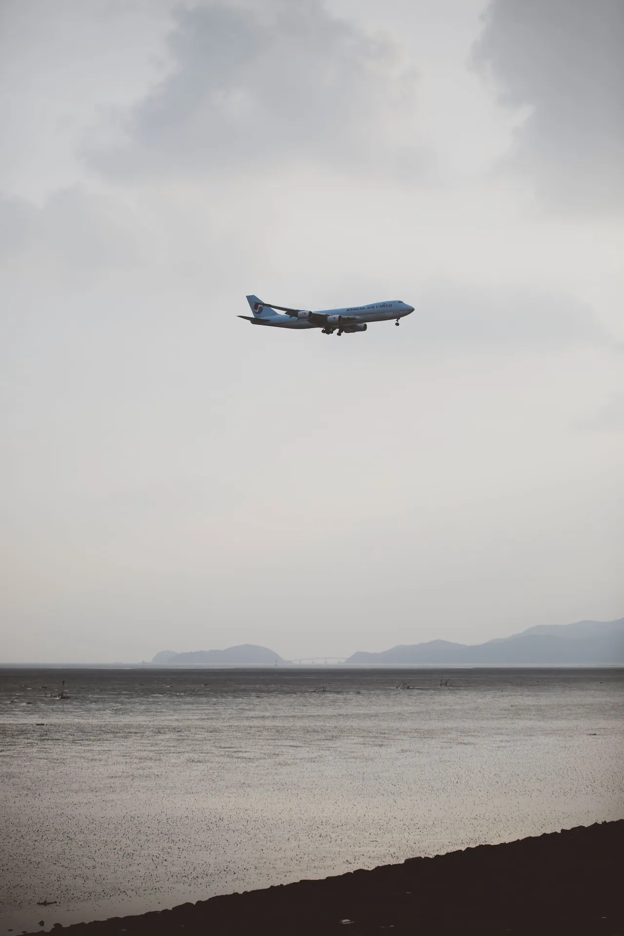 Passenger airplane flying over a calm ocean with distant mountains under a cloudy sky at dusk.
