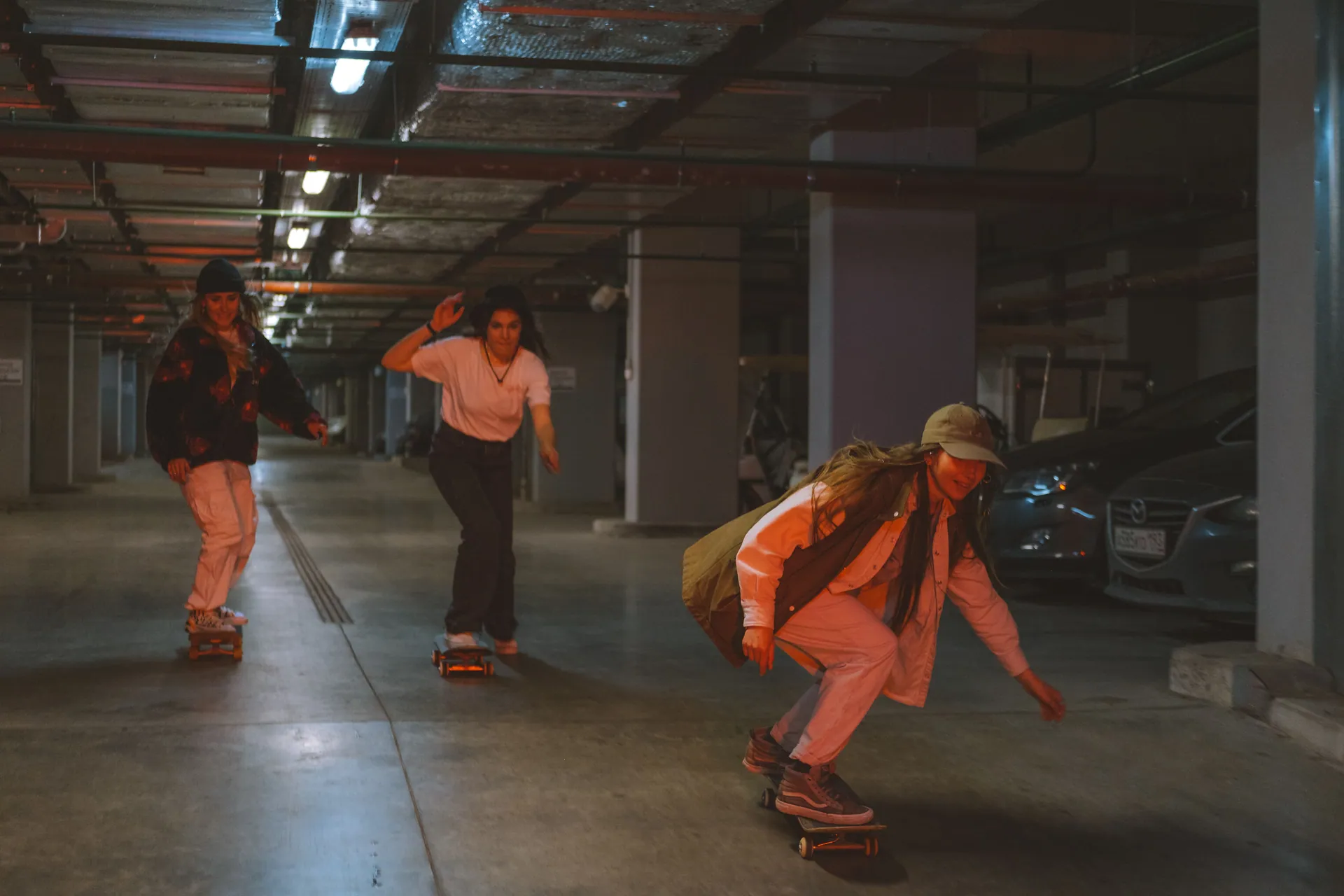 Three young adults skateboarding together in a dimly lit underground parking garage.