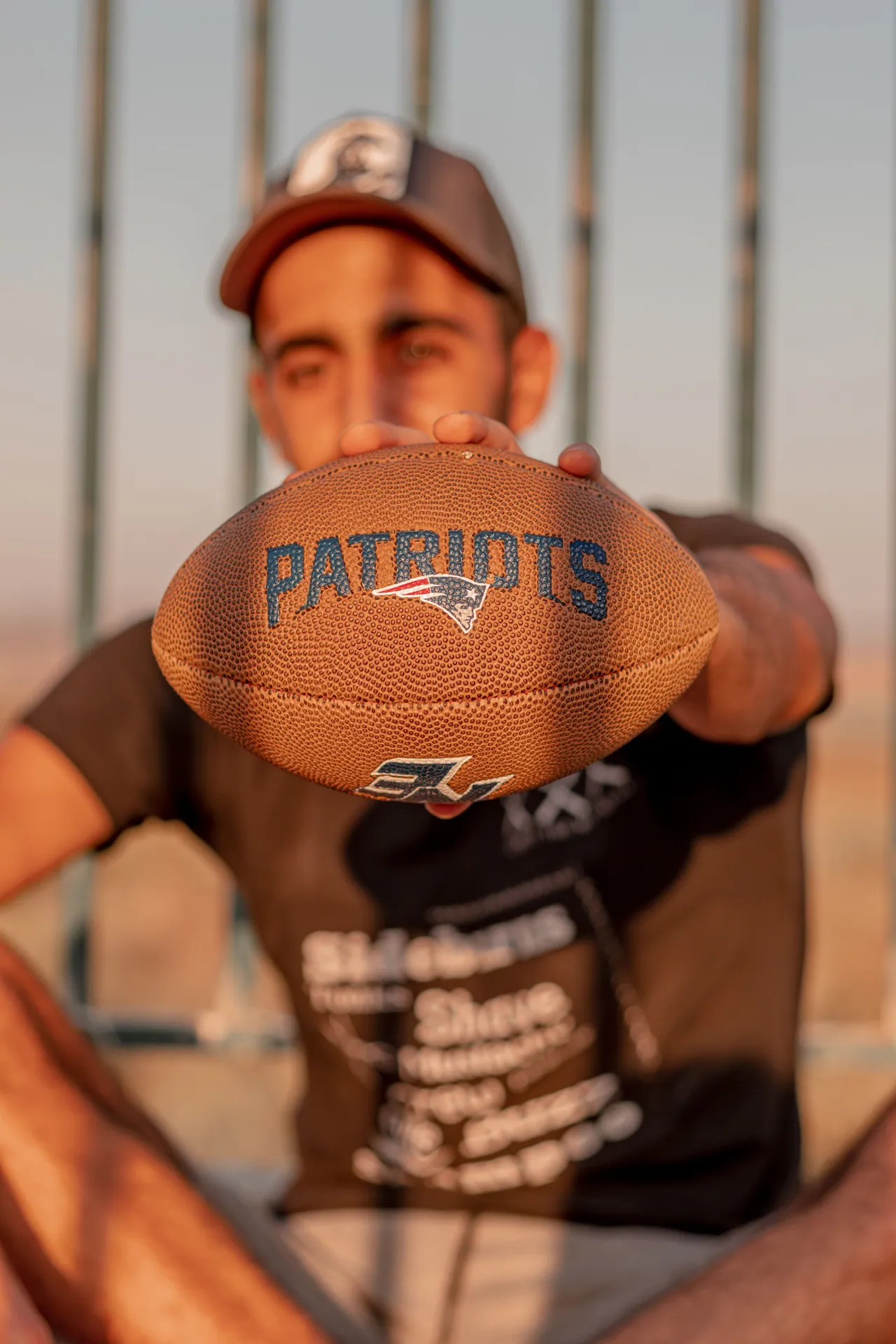 Unrecognizable ethnic male in sportswear and cap sitting near fence and holding brown American soccer ball in hand outside