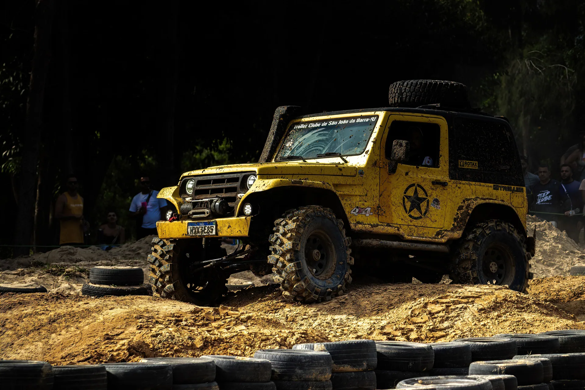 Yellow off-road vehicle navigating through a rugged, muddy terrain during an outdoor challenge event.