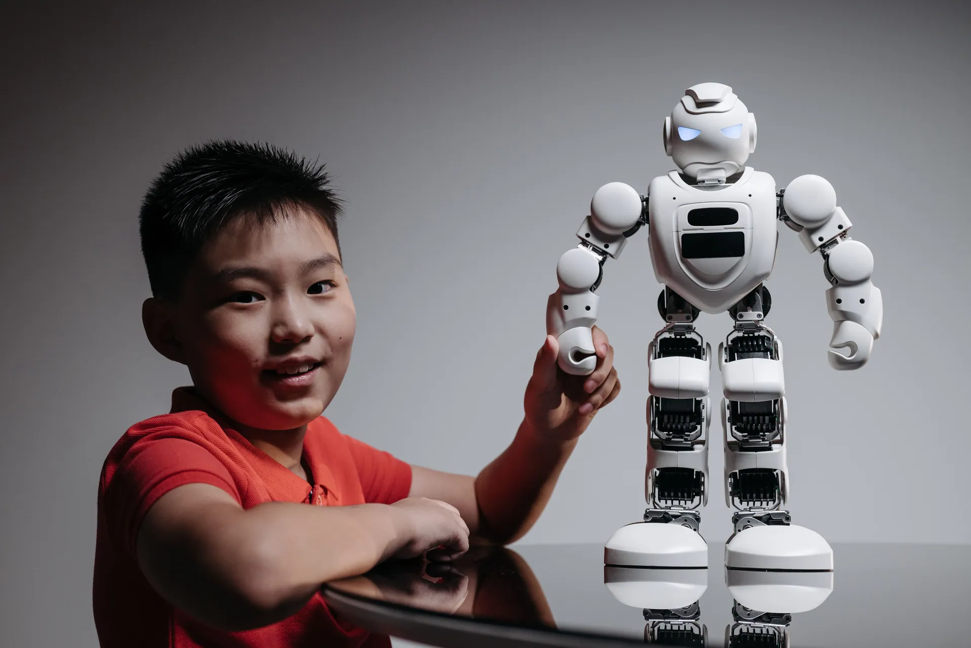 A young boy in a red shirt engaging with a humanoid robot indoors.
