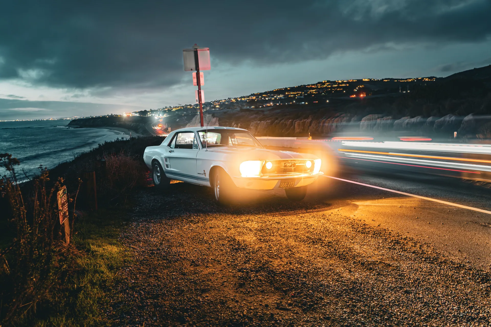 Classic white Ford Mustang with headlights on, parked by Newport Beach coast at sunset.