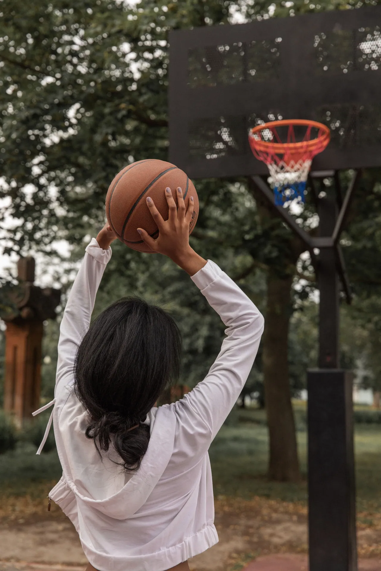 Back view anonymous African American female in white outfit raising arms and throwing basketball ball into hoop on sports ground in green park
