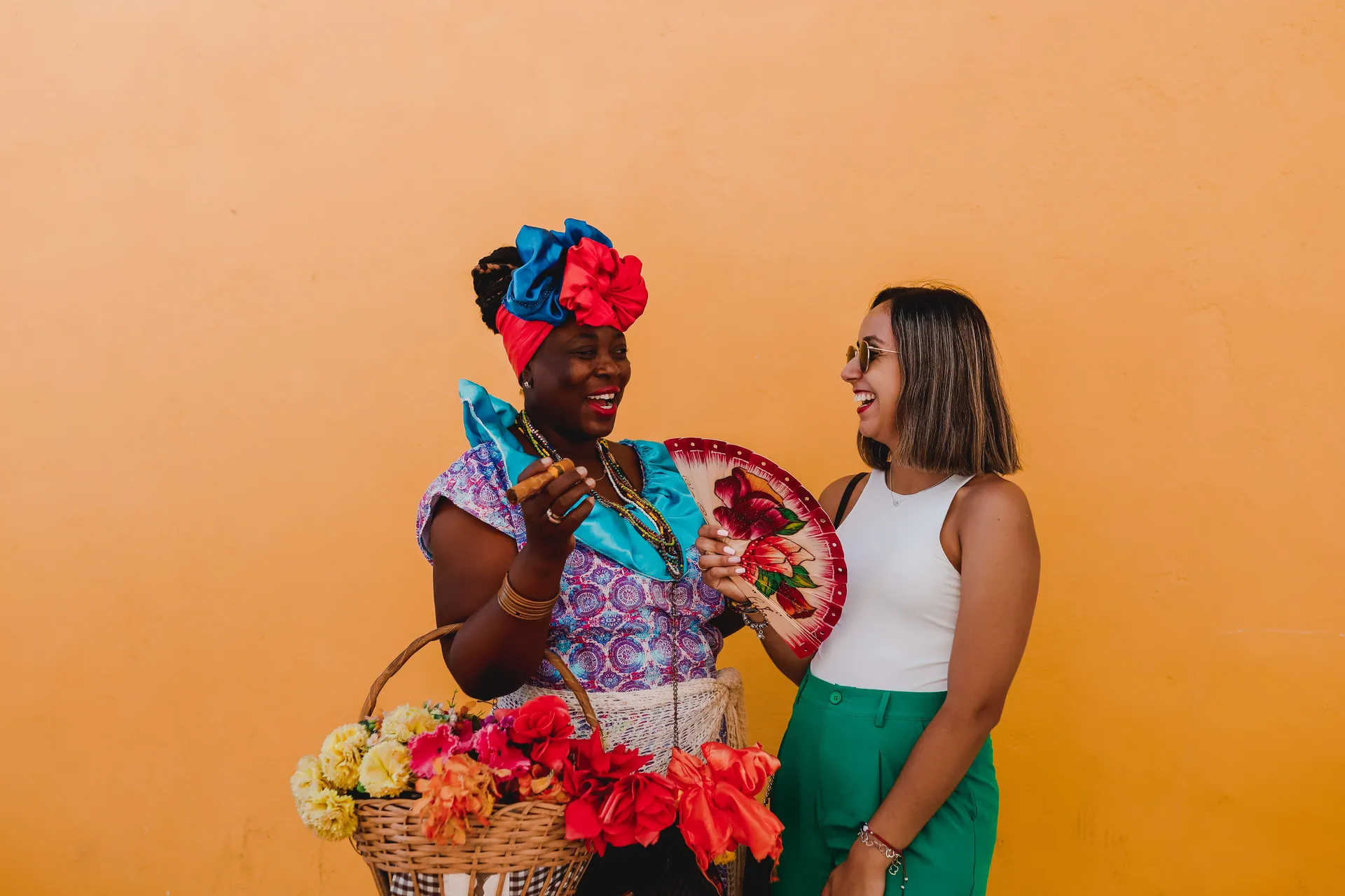 Two women laughing together while holding flowers and a fan against a bright orange background.