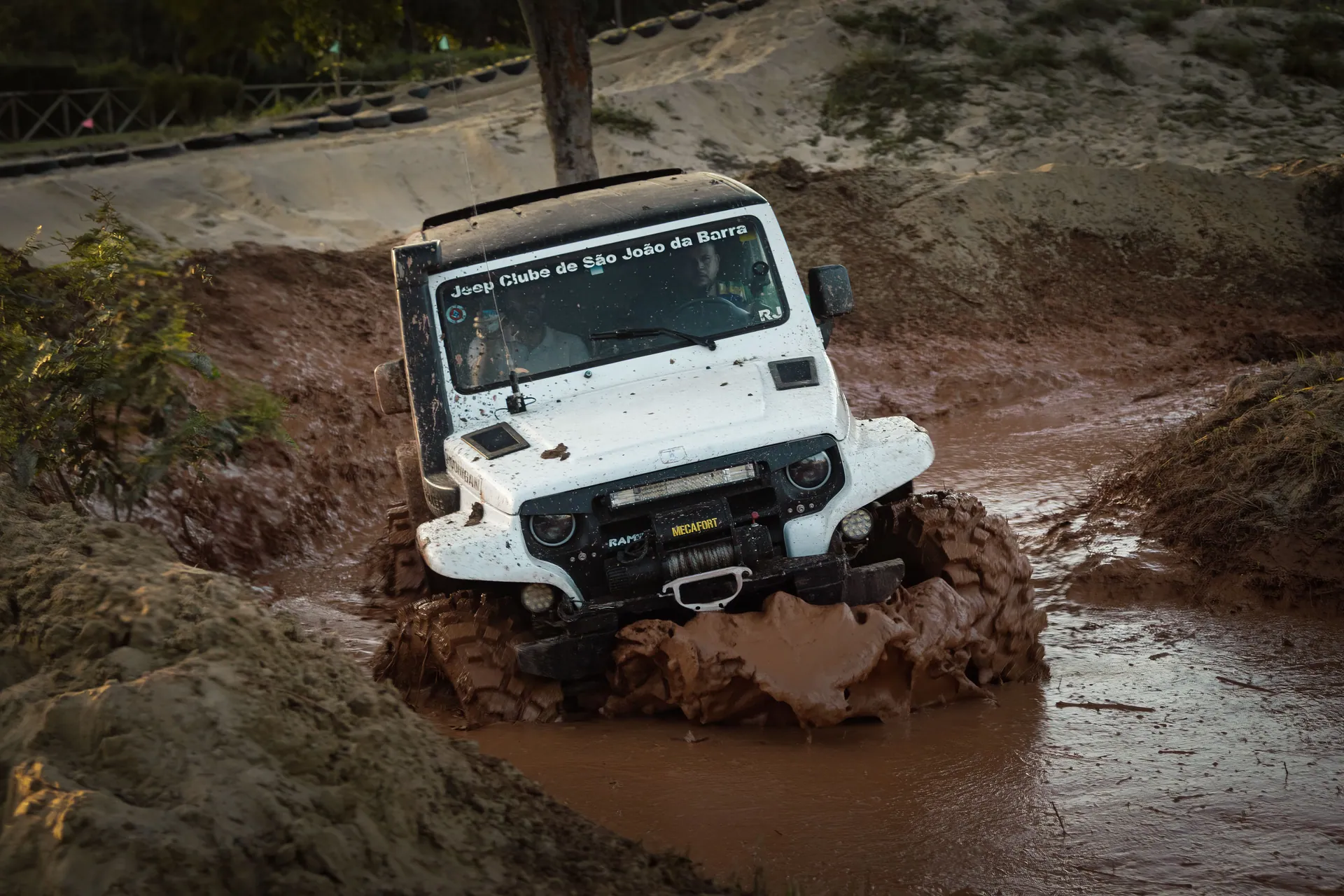 A white jeep navigates through a challenging muddy off-road track during a competition.