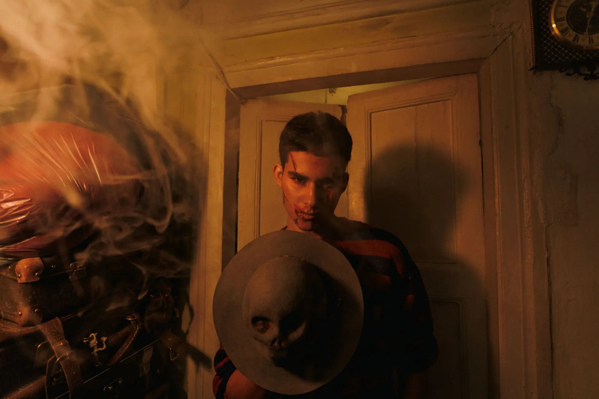 A man in a dimly lit room holds a hat adorned with a skull, surrounded by smoke.