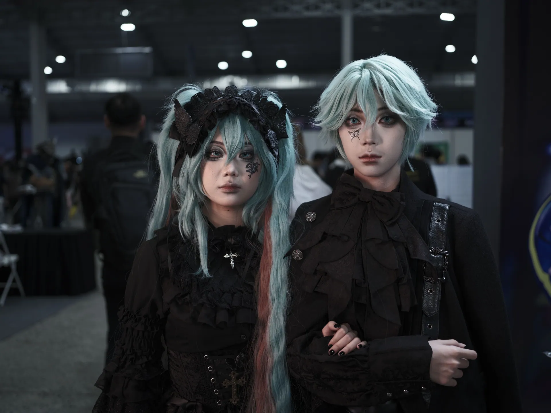 Two individuals in gothic cosplay at an indoor anime convention. Unique and vibrant costumes.