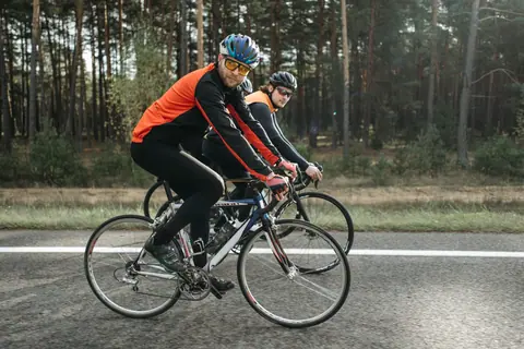 Two adult male cyclists ride on a scenic country road surrounded by a forest backdrop.
