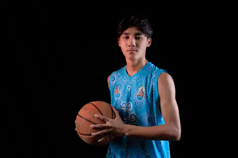 Portrait of a young male basketball player holding a ball against a black background.