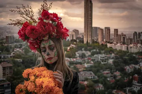 A woman with Day of the Dead makeup holding marigold flowers against Mexico City's skyline.