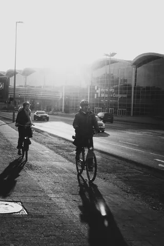 Black and white photo of cyclists riding outside Hamburg Messe in sunlight.