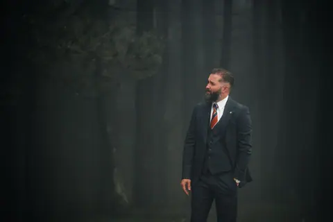 Confident man in a suit stands in a dark, misty forest. Eerie yet professional ambiance.