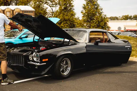 A classic black vintage car with its hood open, parked outdoors, with a person pointing at the engine.