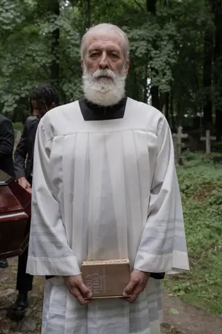An elderly priest holding a bible in a solemn outdoor ceremony, surrounded by nature.