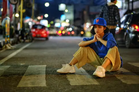 A young woman in casual attire sits on a city street at night, Garut, West Java.
