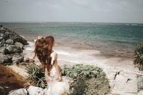 A woman with long hair stands by a rocky seashore, holding a hat in a serene coastal scene.