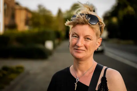 Portrait of a smiling woman outdoors in casual attire, wearing sunglasses and a necklace.