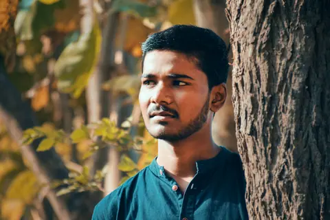 A young man with stubble stands by a tree in a natural setting, looking thoughtful.