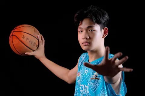 Teenager in blue jersey posing with a basketball against black backdrop.