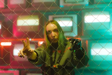 A woman with a prosthetic hand stands behind a chain link fence, bathed in colorful lights.