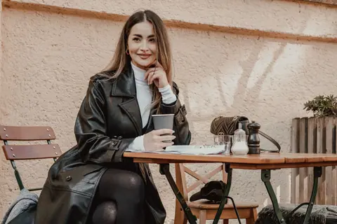 Woman in a black coat smiling while enjoying coffee outdoors at a cafe table.