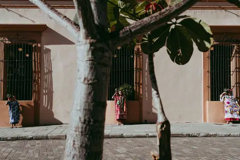 Women in traditional attire standing on a Oaxaca sidewalk, casting shadows in sunlight.