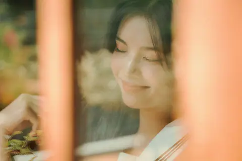 A serene portrait of a young woman taken through a window with a dreamy, soft focus effect.