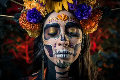 A close-up of a woman with colorful Dia de los Muertos face paint and floral headpiece, reflecting Mexican culture.