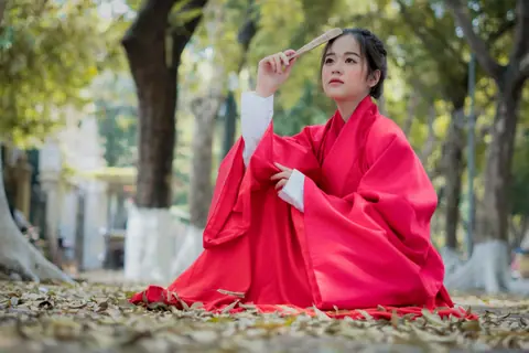 Asian woman in traditional clothing holding a fan, sitting outdoors with a blurred background.