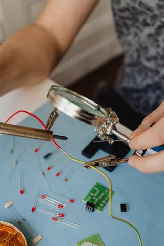 A person soldering wires and components on a work table using a magnifying glass for precision.
