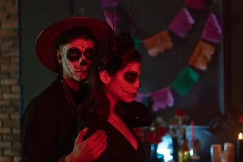 A couple in Día de los Muertos attire and face paint poses with candles and decorations.