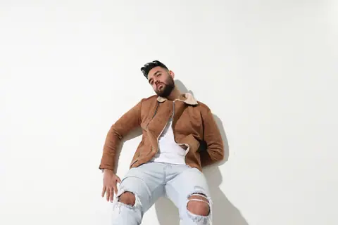 Stylish young man in brown jacket and ripped jeans posing against a white wall.