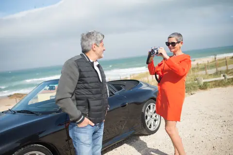 An elderly couple poses with their luxury convertible by the beach in Portugal.