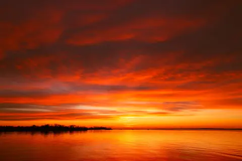 A breathtaking orange and red sunset casting dramatic clouds over a serene lake, offering a moment of tranquility.