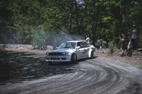 A vintage car drifting around a forest track corner as spectators watch the motorsport event.