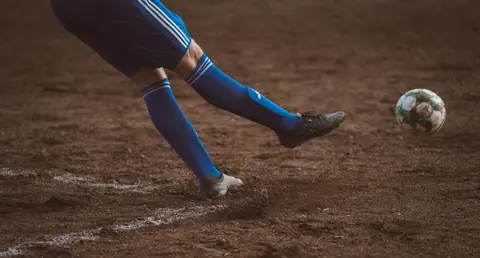 Dynamic soccer player kicks ball on outdoor dirt field. Focus on leg action.