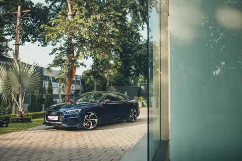 A stylish blue Audi RS5 car parked outdoors on a sunny day with lush greenery.