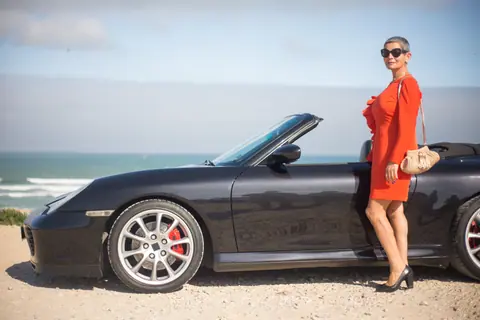 Stylish woman in red dress poses by a luxury black convertible on a scenic beach road.