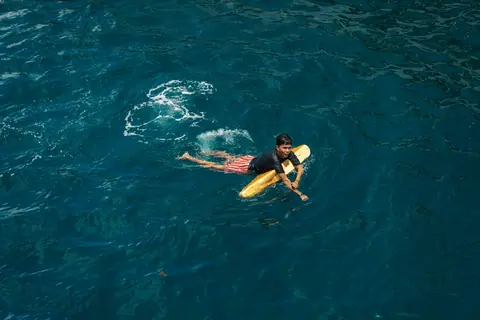A young surfer paddles with a yellow board in clear blue ocean waters.